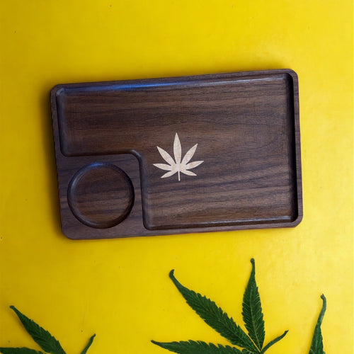 Wooden tray with a cannabis leaf design on a yellow surface with green leaves.
