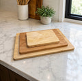 Three nested Maple cutting boards in different wood tones resting on a white marble kitchen countertop near a window.